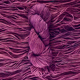 A plummy purple skein of yarn lays vertically atop a bed of the same yarn whose strands are laying horizontally.