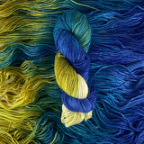 A variegated skein of yarn lays vertically atop a bed of the same yarn.  The yarn is cream, gold, green, teal and deep blue. 
 The bed of yarn has strands laying horizontally.