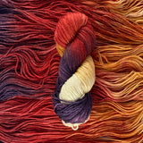 A variegated skein of yarn lays vertically atop a bed of the same yarn.  The yarn is cream, pale orange, deep red, and purple. 
 The bed of yarn has strands laying horizontally.