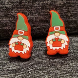 Two silicone sttich markers in the shape of gnomes.  The gnomes are wearing green hats with red belted bands around them.  They are also wearing red trousers and boots.  They are light skinned and have long white beards.  They carry a small red gift box in their hands.  