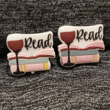 Two silicon stitch stoppers depicting the image of a stack of three books, the topmost of which lays open.  In front of the books is a tall stemmed glass of red wine.  Above the boos and beside the wine glass it says Read in scripted writing.  