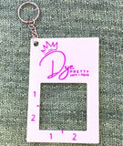 A rectangular, white gauge ruler tool. A keyring comes from the top left corner. The tool lays against a green knitted background.  The tool has the Dye Pretty logo at the top.  A square hole takes up the enter or the tool with 2 inches marked on the left side and on the bottom of the hole. 