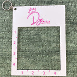 A rectangular, white gauge ruler tool. A keyring comes from the top left corner. The tool lays against a green knitted background.  The tool has the Dye Pretty logo at the top.  A square hole takes up the enter or the tool with 4 inches marked on the left side and on the bottom of the hole. 