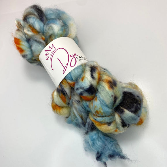 A braid of dyed wool top is cinched at the middle in a white label reading Dye Pretty Yarn + Fibre.  The fibre is dyed blue, orange and black and the braid sits atop a white background.