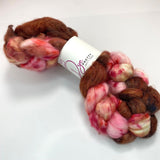 A braid of dyed wool top is cinched at the middle in a white label reading Dye Pretty Yarn + Fibre.  The fibre is dyed pink, red and chocolate brown and the braid sits atop a white background.