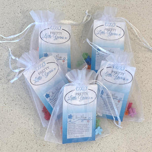 Five white organza bags with ribbon drawstrings lay on a white quartz surface.  The bags contain cards that say Pretty Little Yarns Ltd. in logo form with Clip-a-majig and illegible writing below.  behind the cards you can see some small plastic flower shaped clips.  