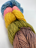 5 mini skeins of worsted weight yarn linked together to resemble a braid.  The skeins are taupey brown, pistachio green, golden yellow, pale pink, and sky blue.  