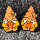 Two silicone stitch stoppers in the form of gnomes. The gnomes are wearing orange hats, mitts and shoes and a golder jumper.  Two small leaves adorn the gnome's hats.  They have white beards and are holding pumpkins in their hands.  