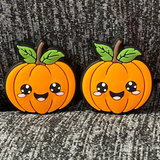 Two silicone stitch stoppers in the form of round pumpkins.  The pumpkins are topped by two leaves and one stem each.  On the front of the pumpkins are smiling kawaii style faces.  