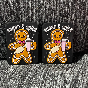 Two silicone stitch stoppers. Rounded black rectangles upon which there are smiling gingerbread people wearing pink bow ties, a cream fanny pack, and carrying a large pink stanley style cup.  above the gingerbread person it reads "sugar & spice" and there are white dots arranged around the gingerbread person on the background.