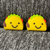 Two silicone stitch stoppers in the form of hard shell tacos filled with meat, cheese, tomatoes, and lettuce.  On the shell, there is a big smiling face.  