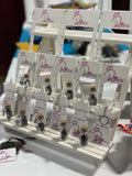 A white painted display stand of 3d printed fibre arts tools and vials of stitch markers.  The bottommost shelf contains 5 glass vials with black stitch markers inside and silver tiaras hanging from the outside. Behind the vials are white wraps per inch tools with dark pink lettering and logo. The next shelf up has more stitch marker vials, and behind the vials is a row of 2"x2" gauge rulers with dark pink Dye Pretty logo.  On the next shelf up is a row of 4"x4" gauge rulers with dark pink Dye Pretty logo. 