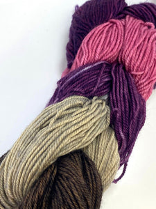 5 mini skeins of fingering weight yarn linked together to resemble a braid.  The skeins are dark grey, light grey, variegated purple and grey, dark pink, and deep purple.  