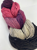 5 mini skeins of worsted weight yarn linked together to resemble a braid.  The skeins are dark grey, light grey, variegated purple and grey, dark pink, and deep purple.  