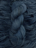 A skein of yarn sits vertically atop a bed of the same yarn laid out with the strands running horizontally.  The yarn is all a mid-tone, tonal denim blue colour.  