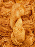 A skein of yarn sits vertically atop a bed of the same yarn laid out with the strands running horizontally.  The yarn is all a tonal peachy orange colour.  