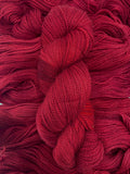 A skein of yarn sits vertically atop a bed of the same yarn laid out with the strands running horizontally.  The yarn is all a deep tonal red colour.  