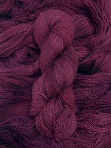 A skein of yarn sits vertically atop a bed of the same yarn laid out with the strands running horizontally.  The yarn is all a tonal deep burgundy/plum colour.  