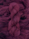 A skein of yarn sits vertically atop a bed of the same yarn laid out with the strands running horizontally.  The yarn is all a tonal deep burgundy/plum colour.  