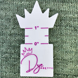 A wraps per inch tool lays against a green knitted background.  It has a tiara shape at the top.  One inch is denoted on the tool and the Dye Pretty logo is on the bottom.  On the reverse, unshown, are the various measurements for the different thicknesses of yarn.  