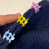 The edges of navy knitted fabric are held together with three plastic flower shaped clips, one light blue, one yellow, and one lilac.  They are being held on a diagonal by a light skinned hand.  