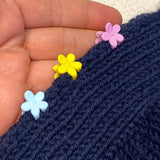 The edges of navy knitted fabric are held together with three plastic flower shaped clips, one light blue, one yellow, and one lilac.  They are being held on a diagonal by a light skinned hand.  